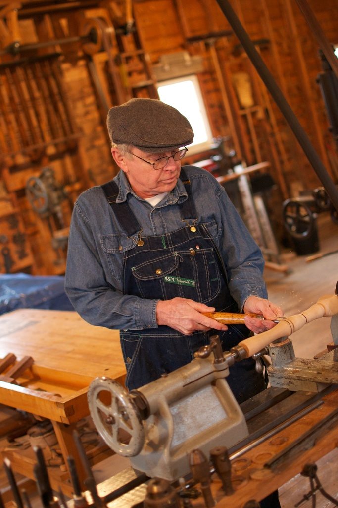A craftsman in a flat cap working at a wooden bench in a workshop